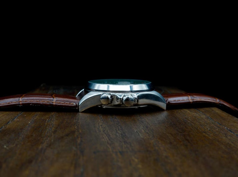 Pu&ograve; includere: Un orologio da polso argentato con cinturino in pelle marrone. Il quadrante dell'orologio &egrave; scuro e il cinturino presenta un motivo strutturato a forma di coccodrillo. L'orologio &egrave; posto su uno sfondo scuro su una superficie di legno.