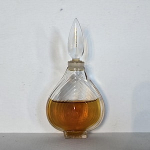 May include: A clear glass perfume bottle with a teardrop-shaped body featuring diagonal ribbed textures. The bottle contains an amber-colored liquid and is topped with a pointed, clear glass stopper that fits into the narrow neck of the vessel.