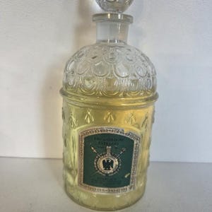 May include: A clear glass perfume bottle with a rounded stopper and a textured surface featuring embossed bee motifs. The bottle contains a pale yellow liquid and has a rectangular green label with the text EAU DE COLOGNE EXTRA DRY.