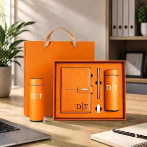 May include: An orange gift set featuring a notebook, a pen, a small USB drive, and two cylindrical vacuum-insulated bottles. Each item is marked with the letters D and T separated by a vertical line. The set is presented in a matching orange box.