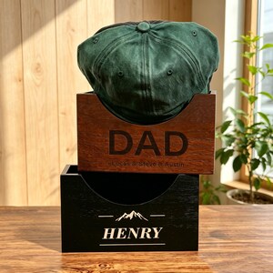 May include: A dark green baseball cap resting on a brown wooden box engraved with the word DAD and the names Lucas & Steve & Austin, stacked atop a black wooden box featuring a mountain graphic and the name HENRY in white lettering.
