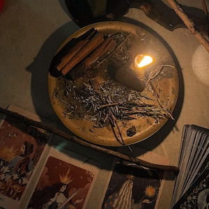May include: A dimly lit scene featuring a brass tray with cinnamon sticks, herbs, and a lit candle. Tarot cards with illustrations and text are scattered around the tray, along with a mirror and wooden sticks. The cards include the text "QUEEN OF SWORDS" and "THE MAGICIAN."
