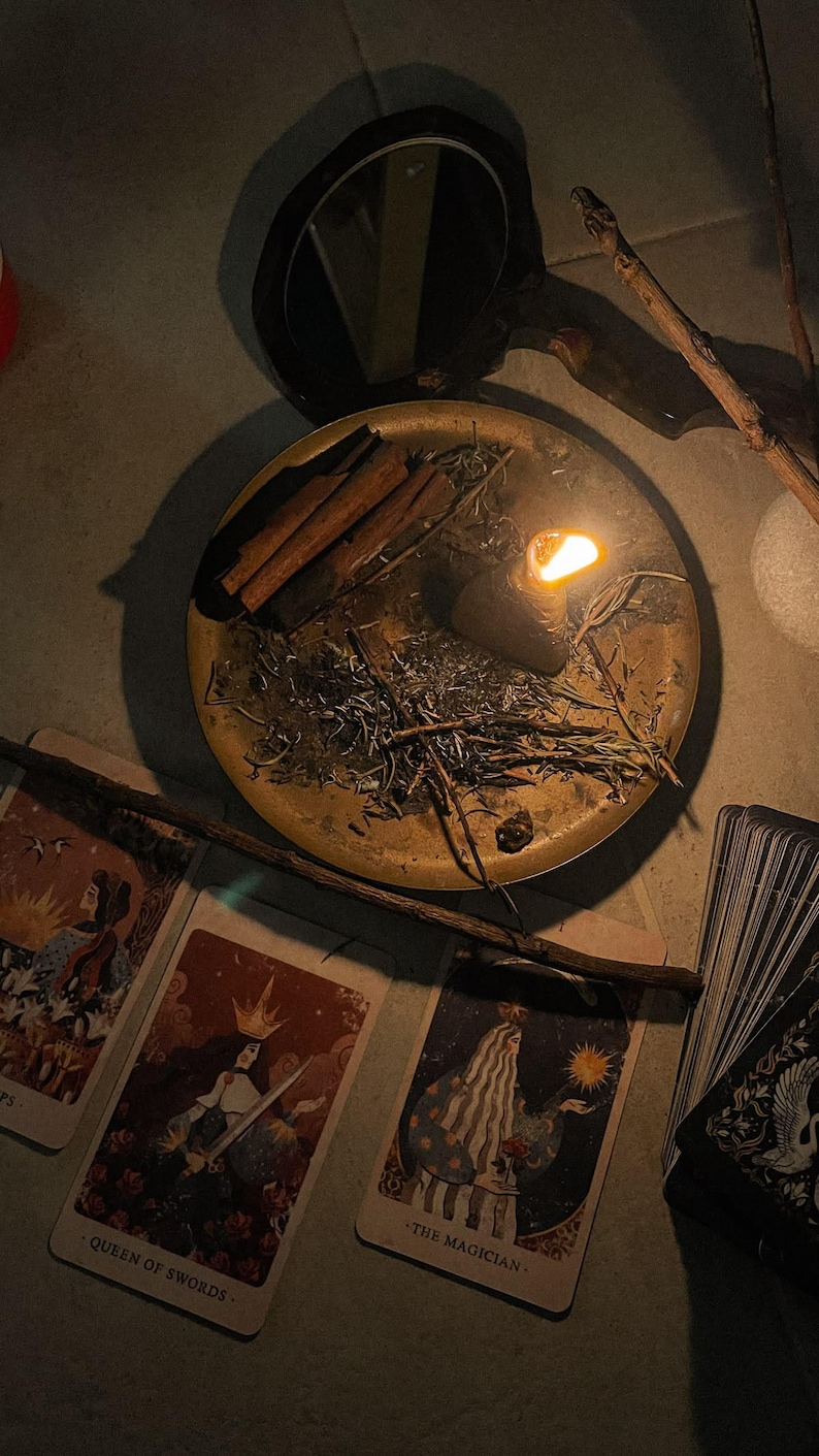 May include: A dimly lit scene featuring a brass tray with cinnamon sticks, herbs, and a lit candle. Tarot cards with illustrations and text are scattered around the tray, along with a mirror and wooden sticks. The cards include the text "QUEEN OF SWORDS" and "THE MAGICIAN."