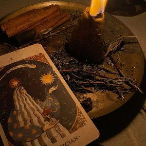 May include: A lit candle with a tall flame sits on a brass plate with a tarot card, cinnamon sticks, and herbs. The tarot card features an illustration of a figure with the text "THE MAGICIAN."