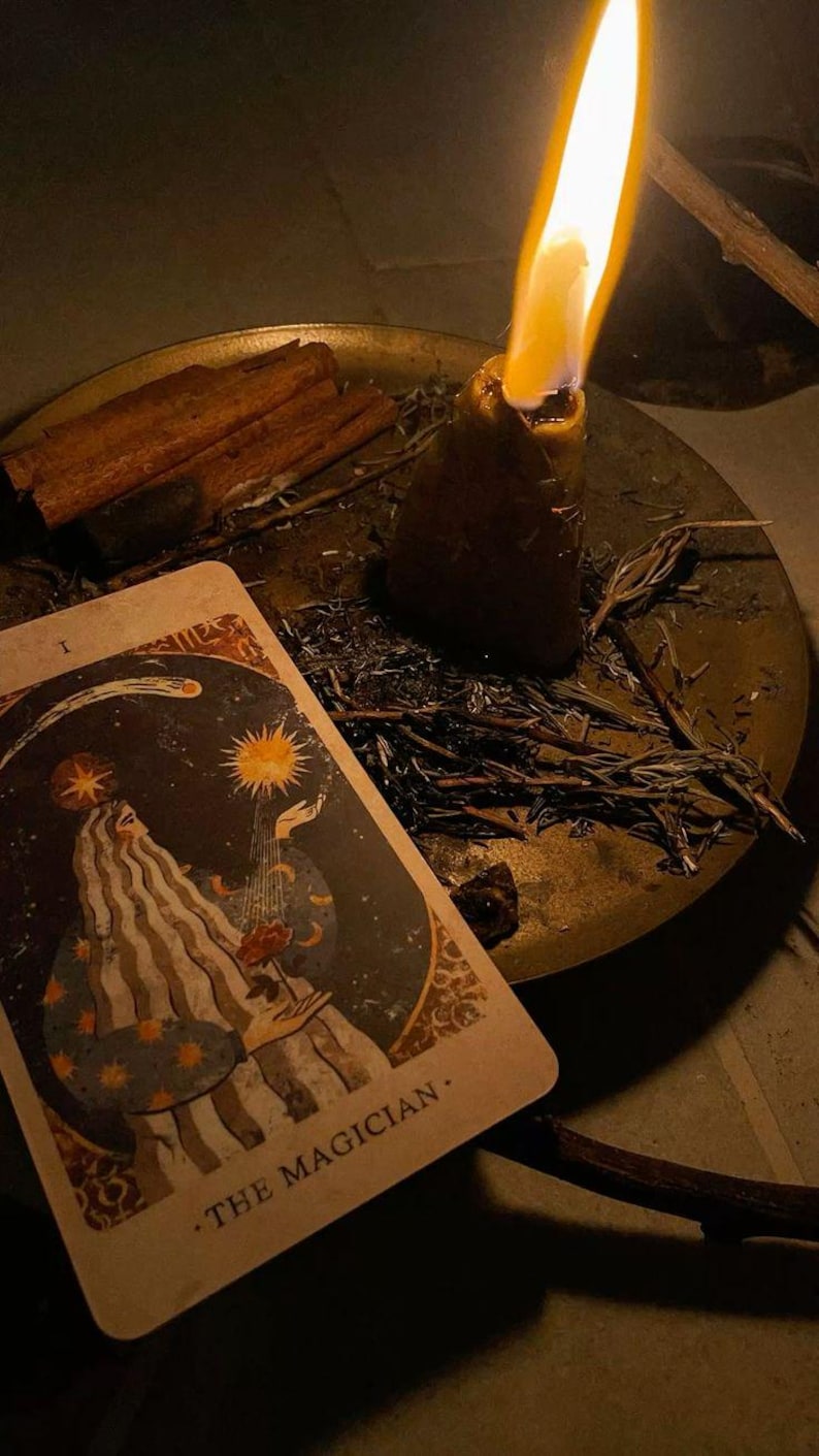 May include: A lit candle with a tall flame sits on a brass plate with a tarot card, cinnamon sticks, and herbs. The tarot card features an illustration of a figure with the text "THE MAGICIAN."