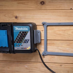 May include: A black and teal Makita DC18C battery charger mounted on a wooden surface. The charger has indicator lights and a blue battery slot. A gray mounting bracket with the word "UP" is attached to the wall.