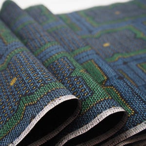 May include: A folded piece of fabric featuring a geometric pattern in shades of blue, green, and black with small yellow accents. The textile has a visible woven texture and a light-colored selvage edge along the bottom of the folds.