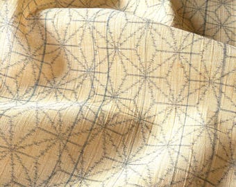 May include: A light yellow fabric featuring a repeating dark grey geometric pattern of intersecting lines and star-like shapes. The material has a visible textured weave with soft folds that create depth and shadows across the surface of the textile.