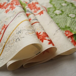May include: A folded length of off-white fabric featuring a repeating pattern of red maple leaves, delicate white cherry blossoms with gray outlines, and a large green textured leaf motif with white floral accents, finished with thin gold lines.