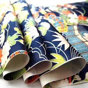 Peut inclure: Un tissu bleu foncé présentant un motif répétitif de fleurs blanches et orange, de feuilles vertes et de formes d'éventails géométriques. Le matériau est plié en plusieurs couches, révélant une texture satinée et un envers clair.