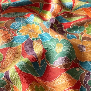 May include: A textured fabric featuring a vibrant floral pattern with a red background. The design includes stylized flowers and leaves in shades of yellow, green, blue, orange, and purple. The material has a distinct crinkled, pebbled surface texture.