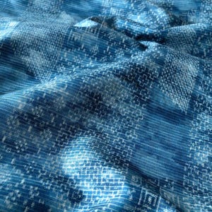 May include: A blue fabric featuring a repeating geometric pattern of white cross-like shapes and linear textures. The material is draped to create soft folds, highlighting the intricate, woven-style design across the surface of the textile.