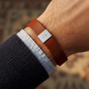 May include: A brown leather bracelet with a silver clasp engraved with the initials "J.S." The bracelet is worn on a wrist over a blue shirt and navy sweater. The leather band is approximately 0.75 inches wide.