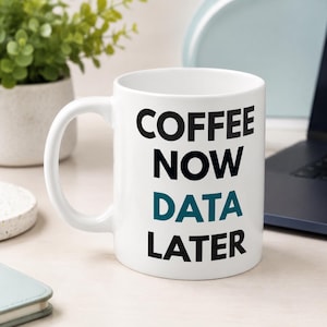 Pode incluir: Uma caneca de cerâmica branca com alça, apresentando o texto preto COFFEE NOW e LATER, com a palavra DATA impressa em azul-petróleo entre eles. A caneca tem um corpo cilíndrico liso e uma alça curva padrão no lado esquerdo para segurar.