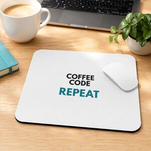 May include: A square white mousepad featuring the black text COFFEE CODE and the teal text REPEAT centred on the surface. A white computer mouse rests on the right side of the mousepad, which has rounded corners and a black edge trim.