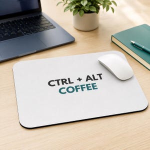 May include: A rectangular white mouse pad featuring the black text CTRL + ALT and the teal text COFFEE printed in the centre. The mouse pad has rounded corners and a black edge, with a white computer mouse resting on its surface.