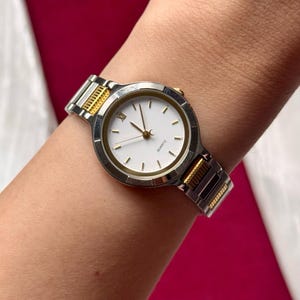 May include: A silver and gold-toned quartz watch with a white face and gold hands. The watch has a silver band with gold accents and is worn on a wrist. The watch face has a minimalist design with gold markers.