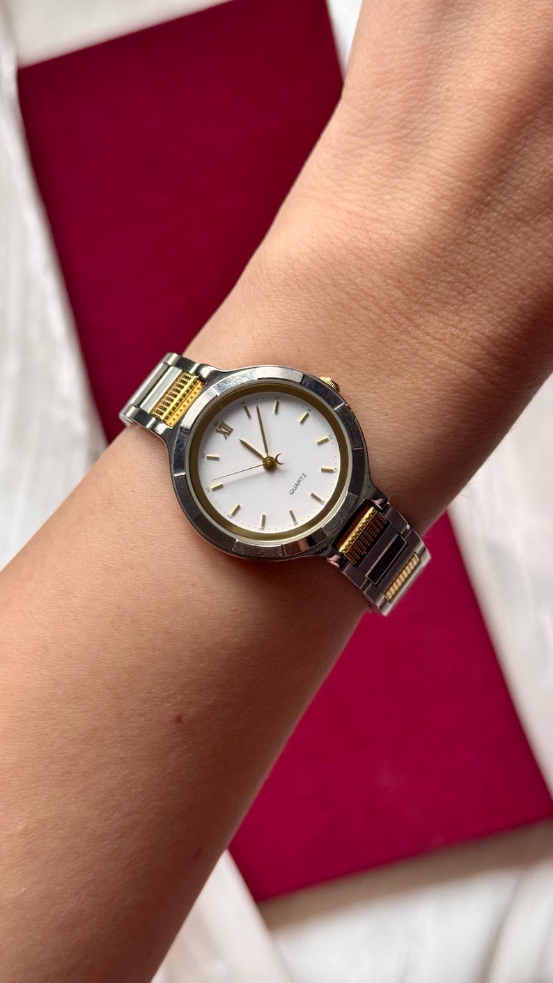 May include: A silver and gold-toned quartz watch with a white face and gold hands. The watch has a silver band with gold accents and is worn on a wrist. The watch face has a minimalist design with gold markers.