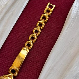 May include: A gold-toned wristwatch with a light-colored face and a decorative band. The band features a series of heart-shaped links. The watch is set against a burgundy background with white fabric.