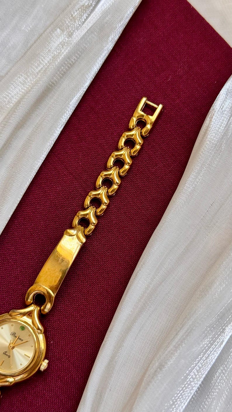 May include: A gold-toned wristwatch with a light-colored face and a decorative band. The band features a series of heart-shaped links. The watch is set against a burgundy background with white fabric.