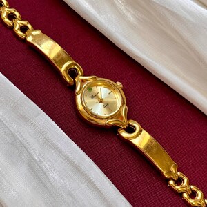 May include: A gold-toned wristwatch with a round face and a linked bracelet. The watch face has a light-colored background with gold hands and markers. The watch is displayed on a burgundy surface with a white fabric draped in the background.