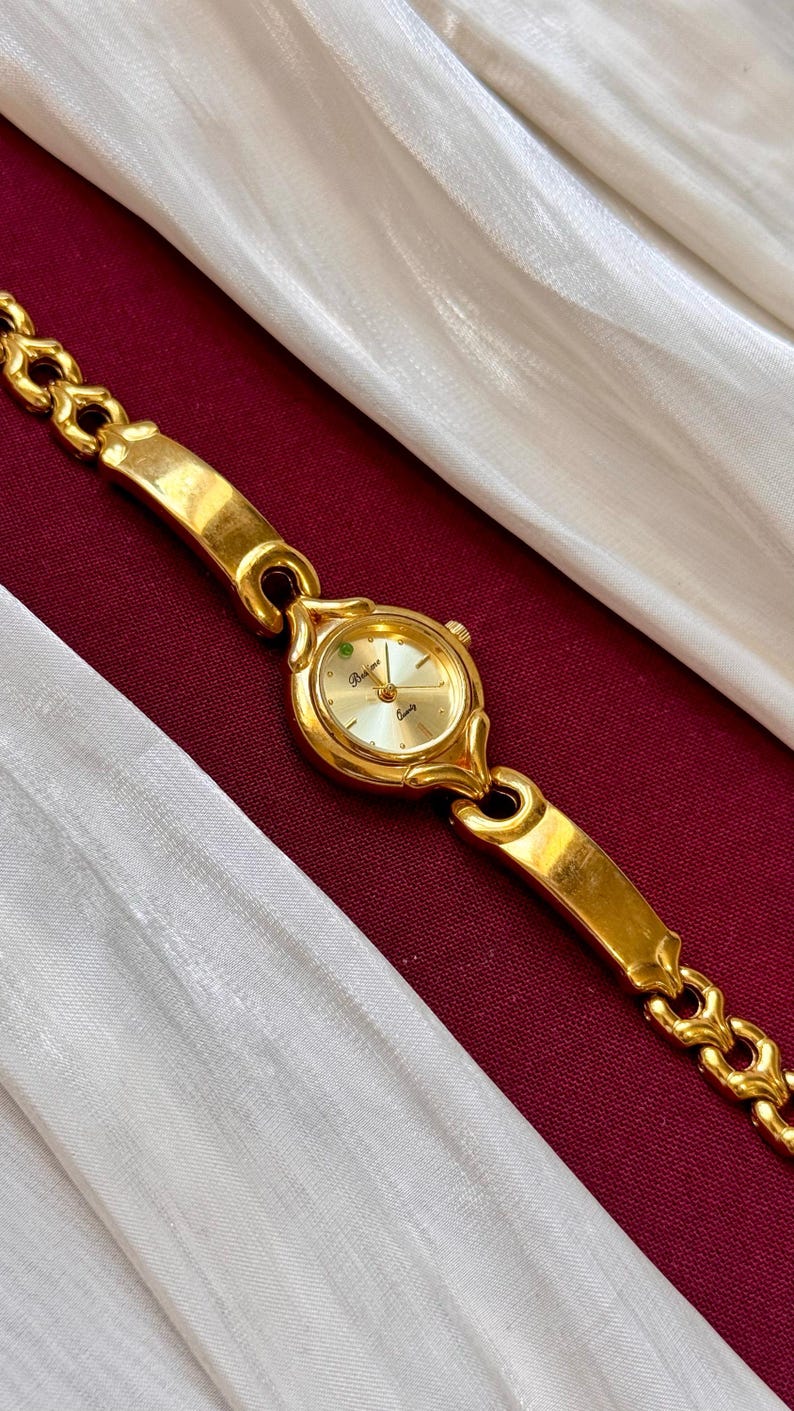 May include: A gold-toned wristwatch with a round face and a linked bracelet. The watch face has a light-colored background with gold hands and markers. The watch is displayed on a burgundy surface with a white fabric draped in the background.