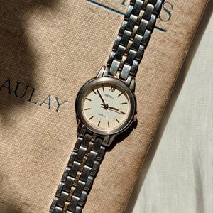 Puede incluir: Un reloj de pulsera Seiko Lucent plateado y dorado con esfera color crema. El reloj tiene una correa de eslabones rectangulares y descansa sobre un libro beige con texto azul. La esfera muestra el logotipo de Seiko y la palabra "Lucent".
