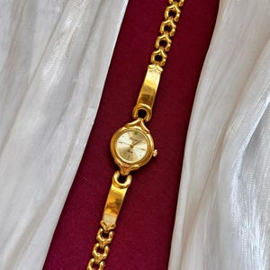 May include: A gold-toned wristwatch with a round face and a linked bracelet. The watch face is a light gold color, and the bracelet has a heart-shaped design. The watch is displayed on a burgundy surface with a white, shimmering fabric.