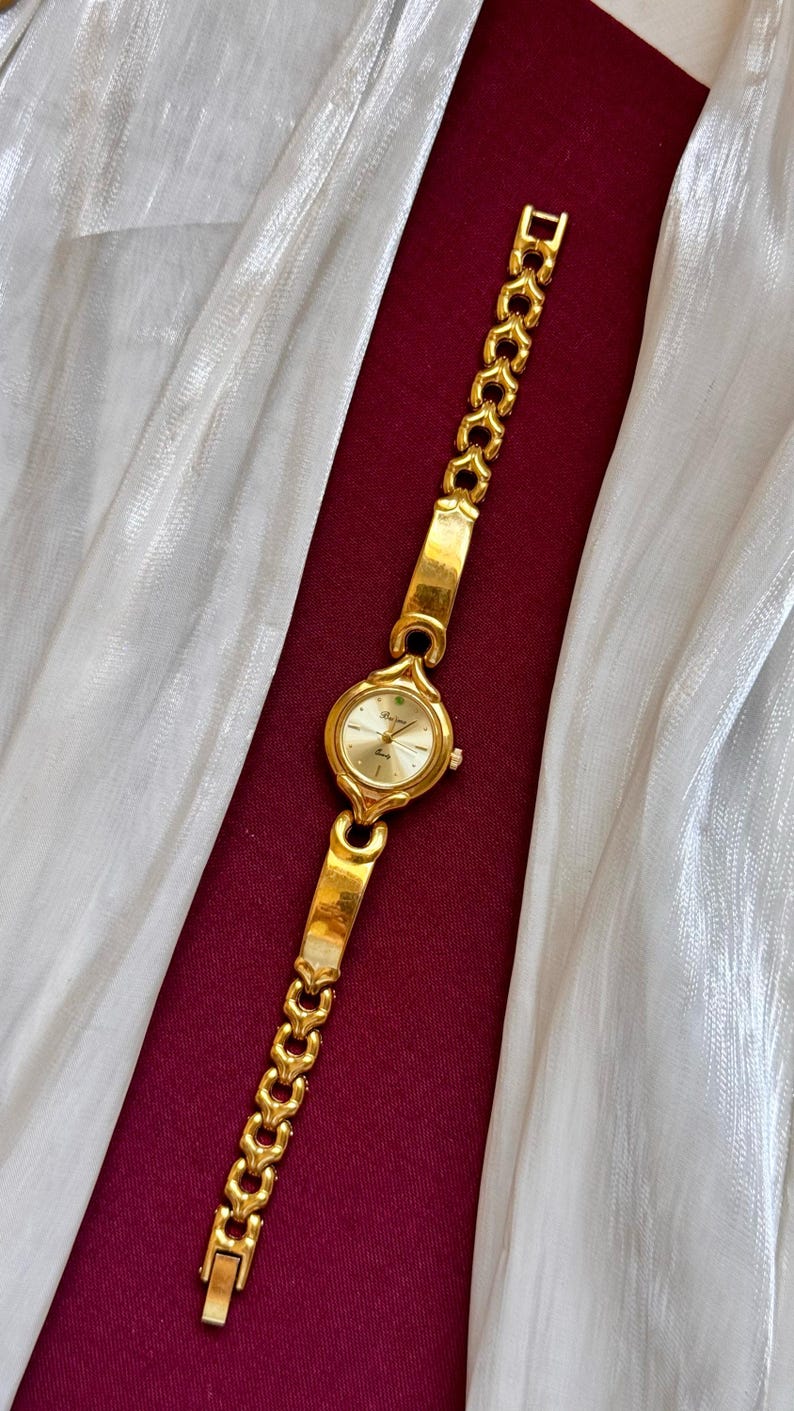 May include: A gold-toned wristwatch with a round face and a linked bracelet. The watch face is a light gold color, and the bracelet has a heart-shaped design. The watch is displayed on a burgundy surface with a white, shimmering fabric.
