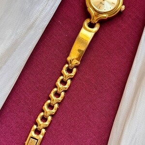May include: A gold-toned wristwatch with a round face and a linked band. The watch face has a gold background with black hands and markers. The band features a series of decorative links. The watch is displayed on a burgundy surface.