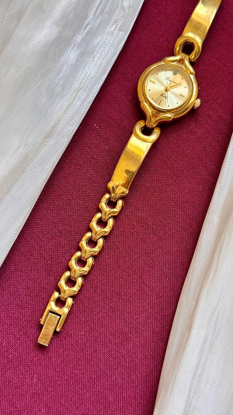 May include: A gold-toned wristwatch with a round face and a linked band. The watch face has a gold background with black hands and markers. The band features a series of decorative links. The watch is displayed on a burgundy surface.