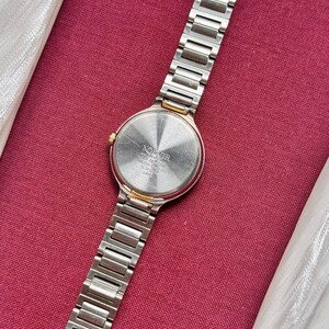 May include: A silver-toned wristwatch with a stainless steel band and a round face. The watch has a gold-colored bezel and a silver-colored dial. The watch is displayed on a red surface.