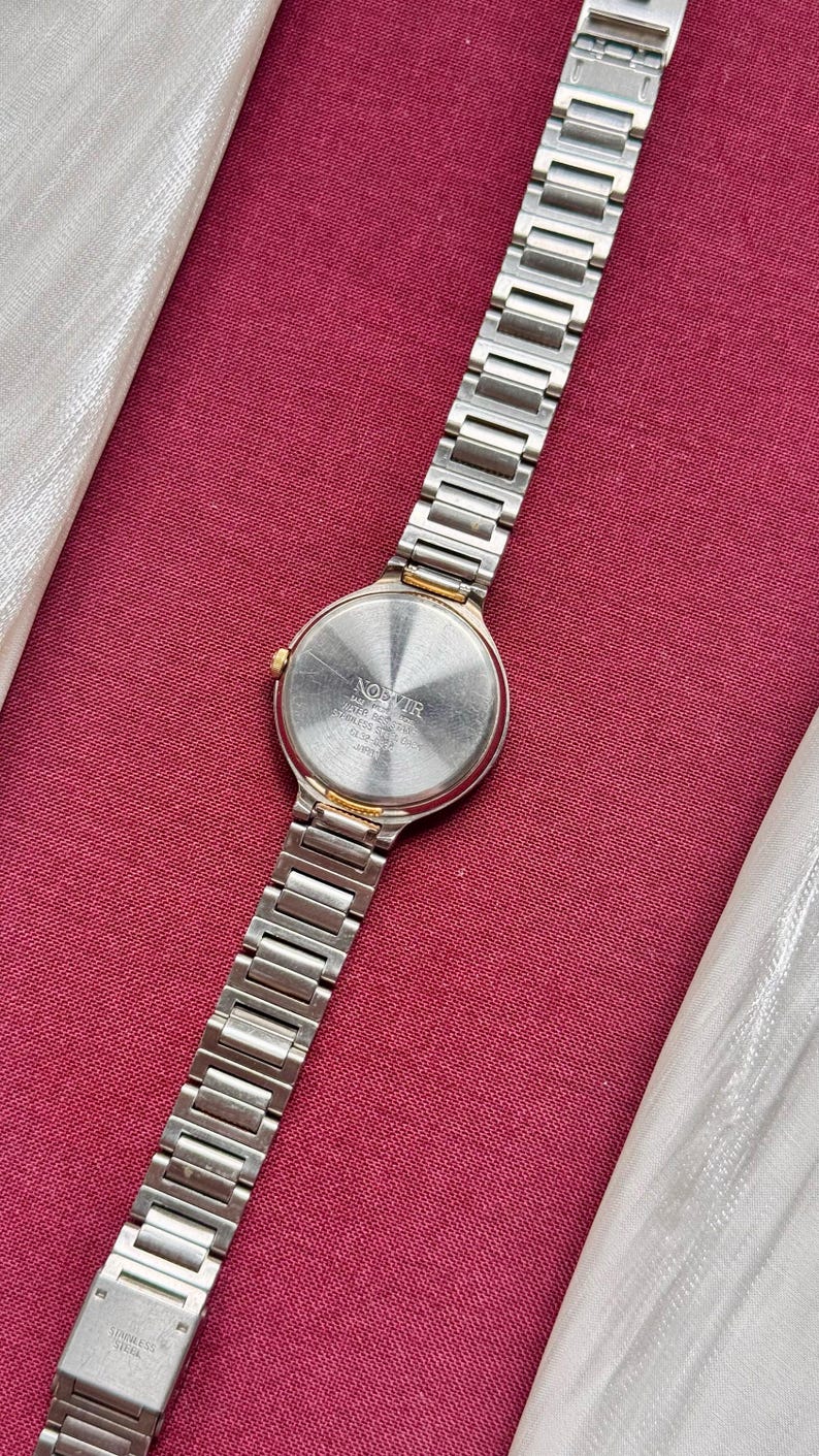 May include: A silver-toned wristwatch with a stainless steel band and a round face. The watch has a gold-colored bezel and a silver-colored dial. The watch is displayed on a red surface.
