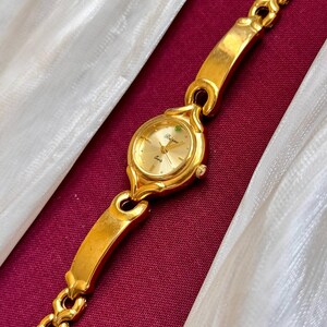 May include: Gold-toned wristwatch with a gold band. The watch face is round with gold hands and a small green gem. The band is a chain link style. The watch is displayed on a burgundy surface with white fabric.