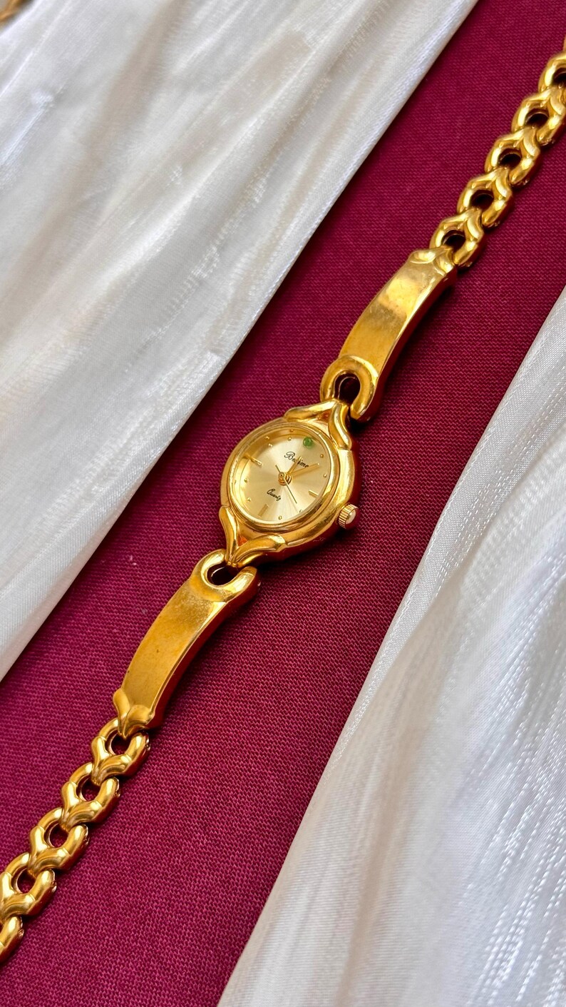 May include: Gold-toned wristwatch with a gold band. The watch face is round with gold hands and a small green gem. The band is a chain link style. The watch is displayed on a burgundy surface with white fabric.
