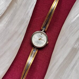 May include: A vintage Alba quartz watch with a silver and gold-tone bracelet. The watch face is white with silver hands and markers. The watch has blue accents and is set against a red and white background.