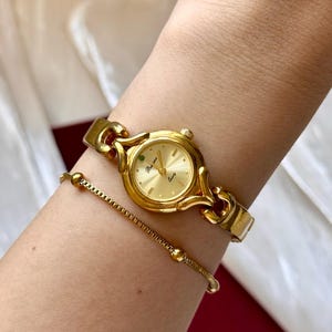 May include: A gold-toned wristwatch and bracelet set. The watch features a round face with a gold dial and a green accent. The bracelet is a delicate chain with small, round beads. The watch and bracelet are worn on a wrist.