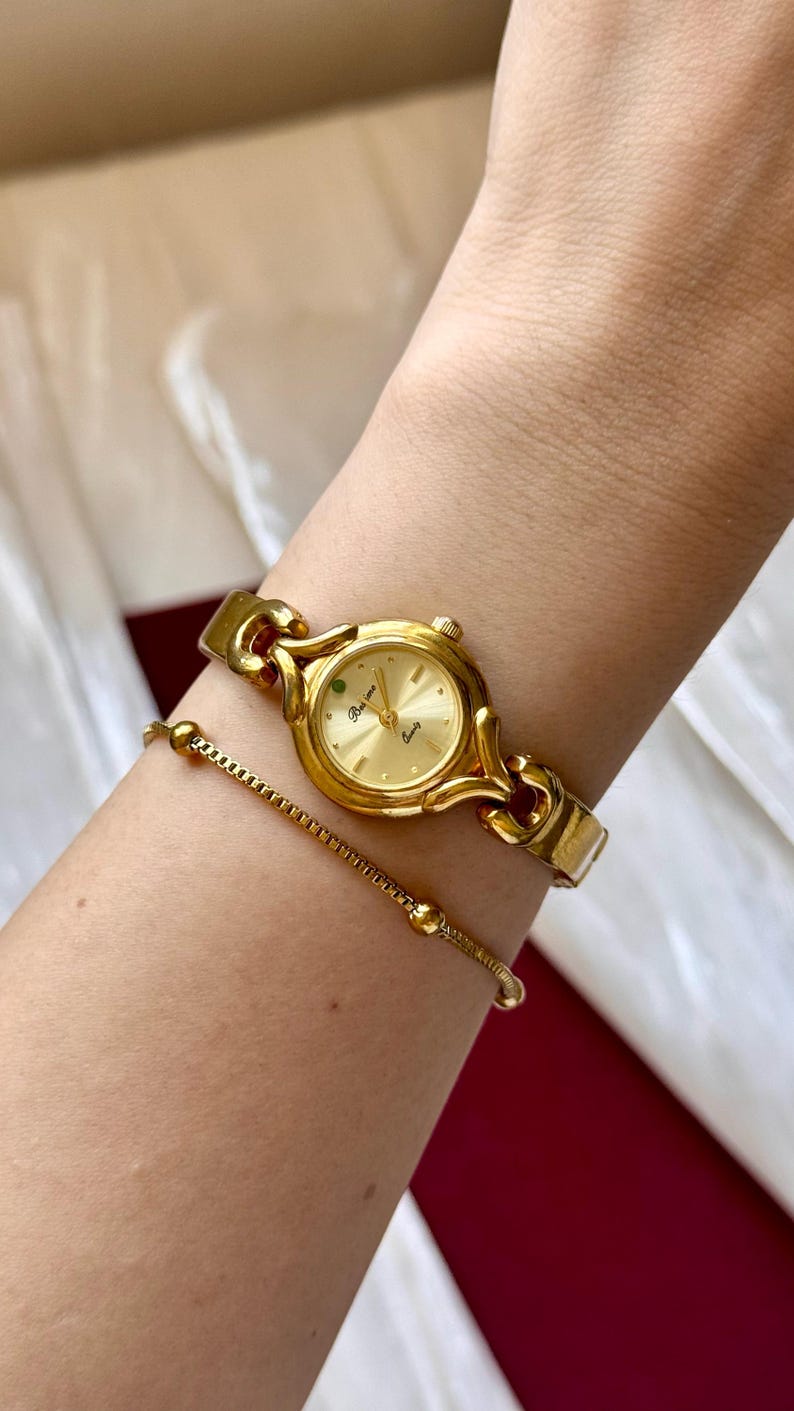 May include: A gold-toned wristwatch and bracelet set. The watch features a round face with a gold dial and a green accent. The bracelet is a delicate chain with small, round beads. The watch and bracelet are worn on a wrist.