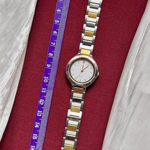 May include: A silver and gold-toned wristwatch with a white face and Roman numeral hour markers. The watch is next to a purple ruler with metric measurements. The watch band is made of silver and gold links.