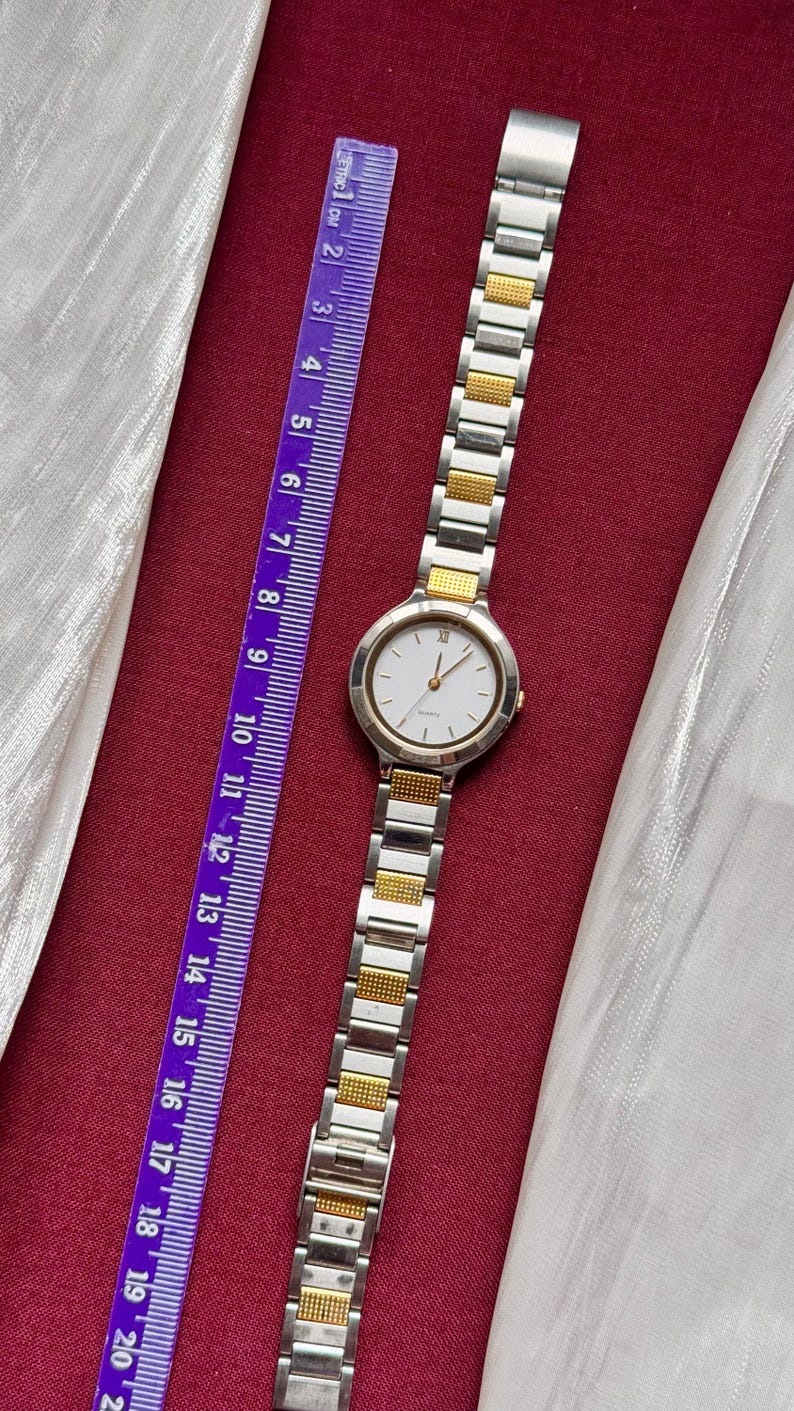 May include: A silver and gold-toned wristwatch with a white face and Roman numeral hour markers. The watch is next to a purple ruler with metric measurements. The watch band is made of silver and gold links.
