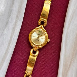 May include: Gold-toned wristwatch with a round face and a matching link bracelet. The watch face is light gold with black hands and the brand name "Barjona". The watch is displayed on a burgundy surface with a white fabric.