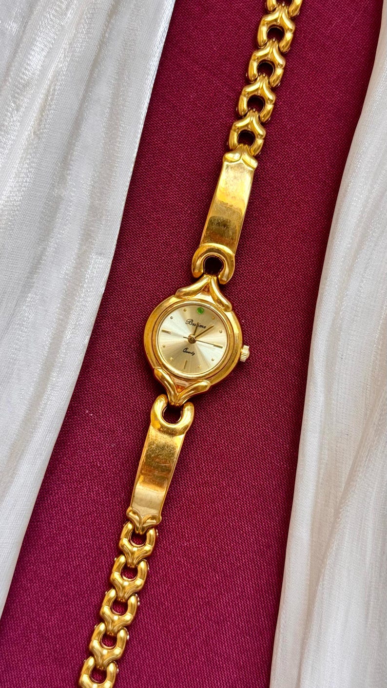 May include: Gold-toned wristwatch with a round face and a matching link bracelet. The watch face is light gold with black hands and the brand name "Barjona". The watch is displayed on a burgundy surface with a white fabric.