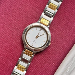 May include: A silver and gold-toned quartz watch with a white face and gold hands. The watch has a round face with Roman numeral XII and gold accents. The watch band is a combination of silver and gold links.