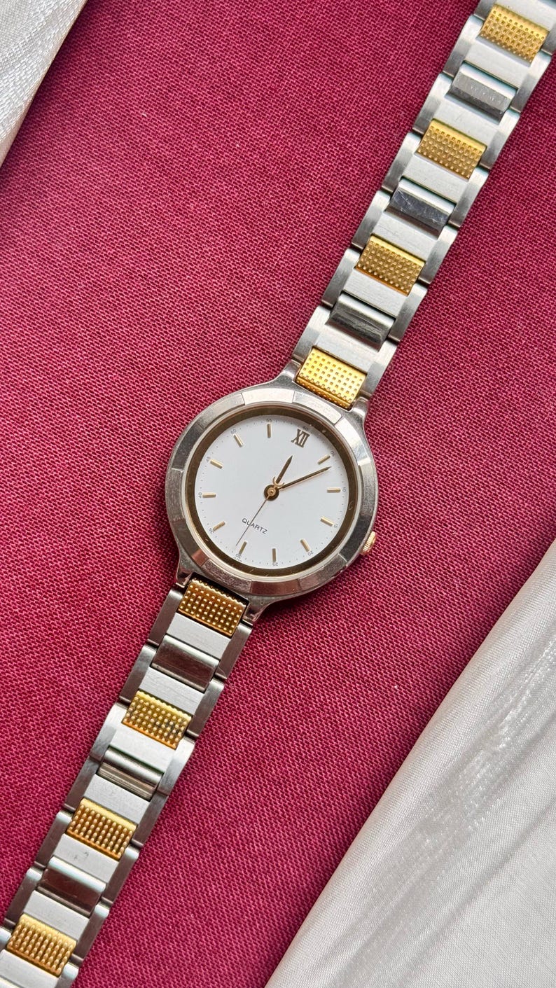 May include: A silver and gold-toned quartz watch with a white face and gold hands. The watch has a round face with Roman numeral XII and gold accents. The watch band is a combination of silver and gold links.