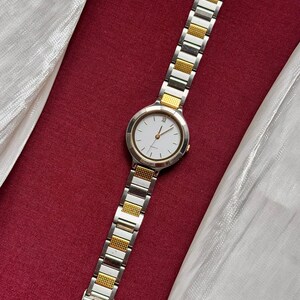 May include: A silver and gold-toned wristwatch with a white watch face. The watch has a rectangular link band with gold-toned accents. The watch is displayed on a red surface with a white fabric.