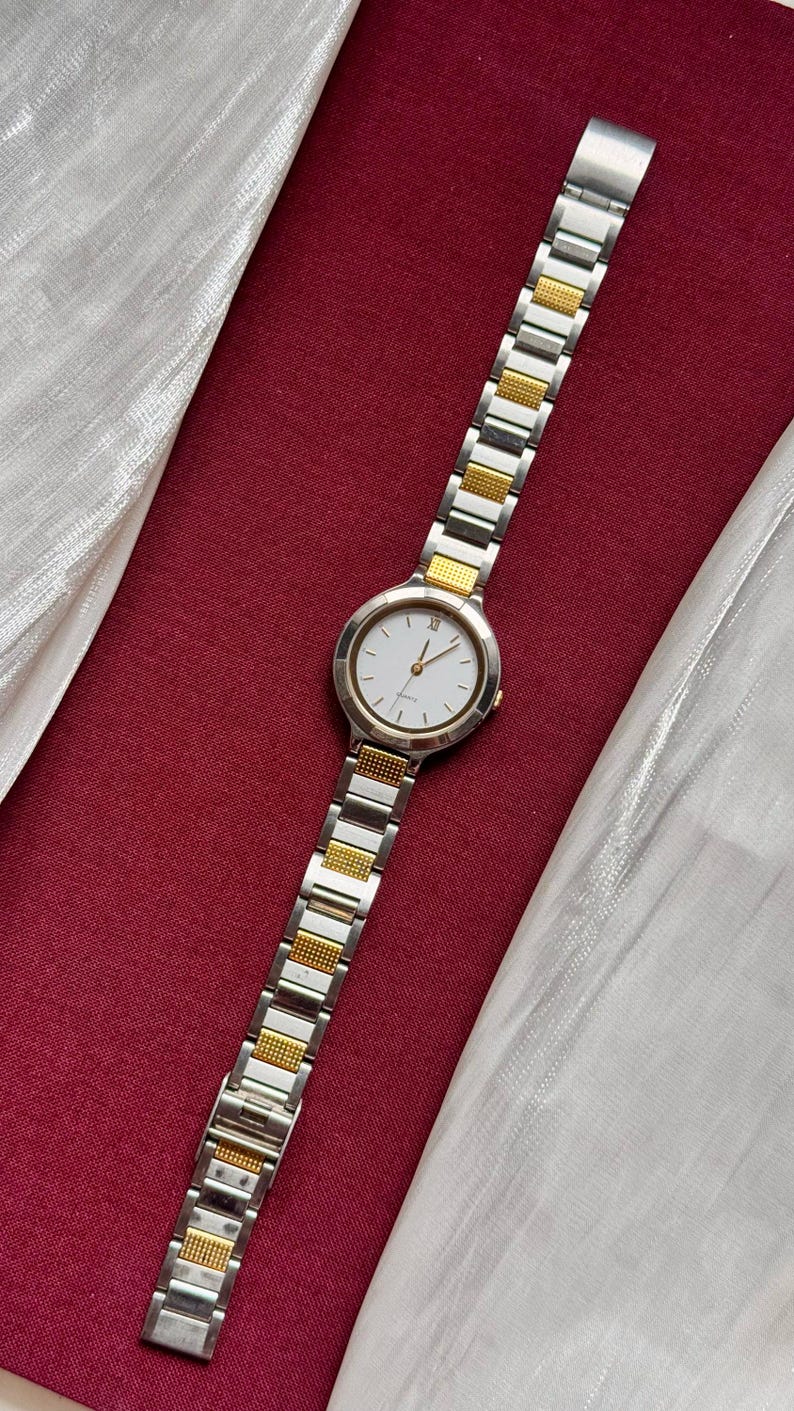 May include: A silver and gold-toned wristwatch with a white watch face. The watch has a rectangular link band with gold-toned accents. The watch is displayed on a red surface with a white fabric.