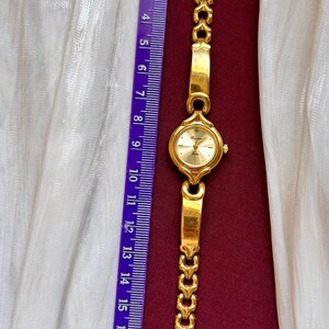May include: A gold-toned wristwatch with a round face and a linked band. The watch face is light gold with black numerals. The band is approximately 7 inches long. A purple ruler is next to the watch.