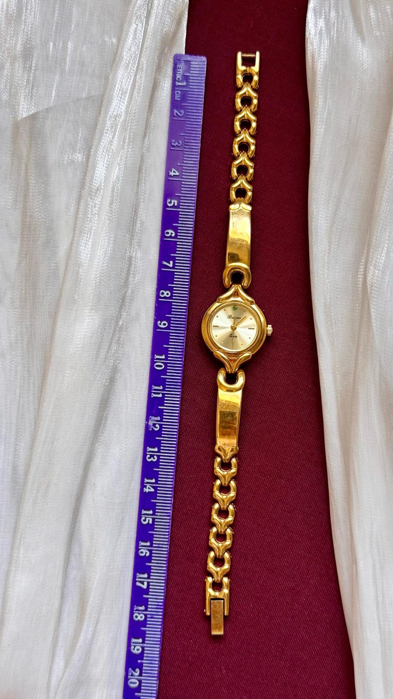 May include: A gold-toned wristwatch with a round face and a linked band. The watch face is light gold with black numerals. The band is approximately 7 inches long. A purple ruler is next to the watch.