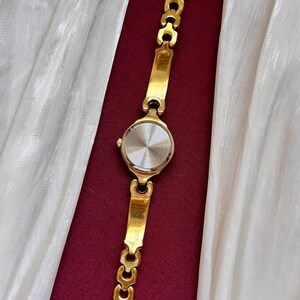 May include: A gold-toned wristwatch with a round face and a linked bracelet. The watch is displayed against a backdrop of white fabric and a deep red surface. The watch band is approximately 7 inches long.