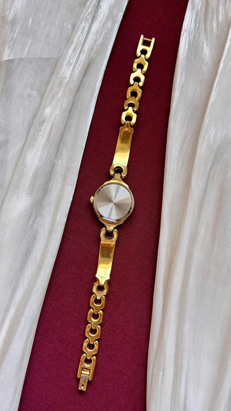 May include: A gold-toned wristwatch with a round face and a linked bracelet. The watch is displayed against a backdrop of white fabric and a deep red surface. The watch band is approximately 7 inches long.